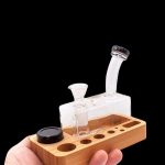 A hand holds a wooden dab station with a glass oil rig and accessories against a black background.