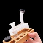 A hand holds a wooden rolling tray with a small glass water pipe (bong) and containers for cannabis concentrates.