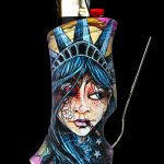 A lighter sleeve depicting a stylized, haunting portrait of Lady Liberty with cannabis leaves and stars, paired with a metal dab tool against a black background.