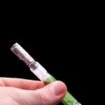 Hand holding a glass cannabis pipe with green and clear accents against a black background.
