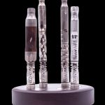 Five glass cannabis oil vape cartridges displayed on a black circular stand, against a dark background.