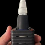 A hand holds a black and orange cannabis vaporizer with a glass mouthpiece.
