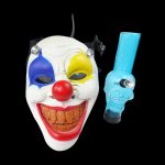 Creepy clown mask with a blue glass pipe positioned beside it on a black background.