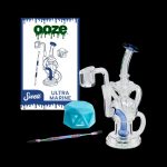A studio shot of a clear glass water pipe ("rig") alongside cannabis accessories including a dab tool, silicone container, and product packaging for "Ooze Swell Ultra Marine."