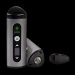 Silver and black cannabis vaporizer with illuminated green controls, displayed with open chamber.