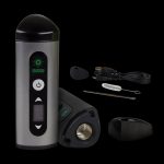 Silver cannabis vaporizer pen with included mouthpiece, cleaning tools, and charging base displayed on a black background.