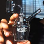 A hand holds a clear glass dab rig with the brand “GRAV” printed on the side, with a separate glass carb cap blurred in the background.