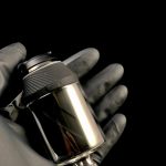 A gloved hand holds a sleek, metallic cannabis oil rig with a glass chamber.