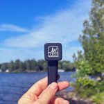 A hand holds a sleek black cannabis vape pen with a white "JW Jane West" logo against a blurred lakeside and blue sky backdrop.