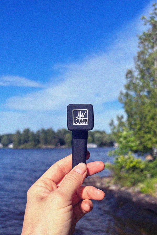 A hand holds a sleek black cannabis vape pen with a white "JW Jane West" logo against a blurred lakeside and blue sky backdrop.