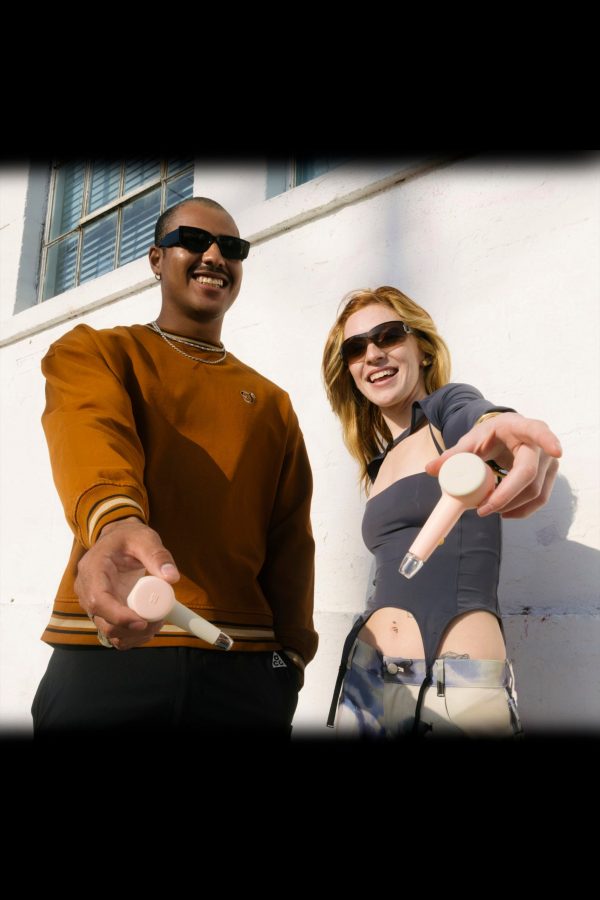 Two people playfully extend lit joints towards the viewer, wearing casual streetwear and sunglasses, against a textured wall.