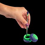 A hand holding a dab tool over a silicone container, both with iridescent purple and green coloring.