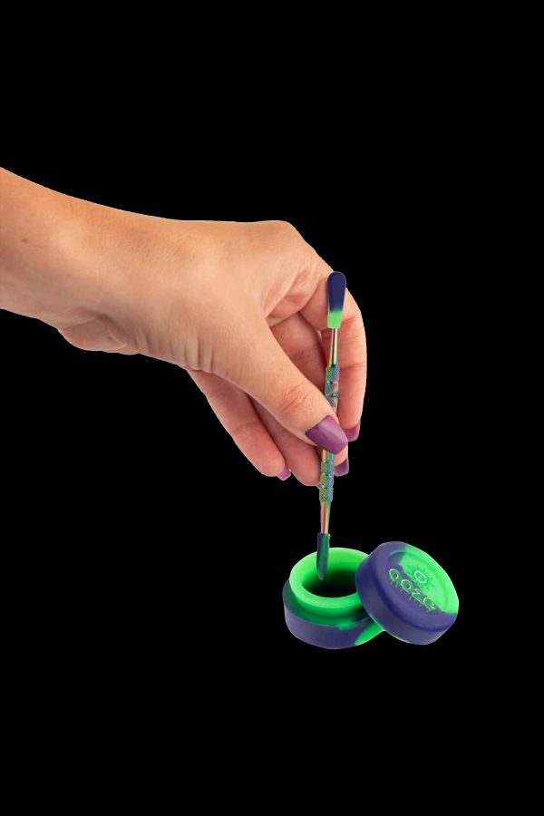 A hand holding a dab tool over a silicone container, both with iridescent purple and green coloring.