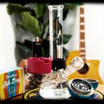 A glass bong with a pink accent piece, lighter, grinder, and ashtray on a patterned surface, with a blurred plant and guitar in the background.