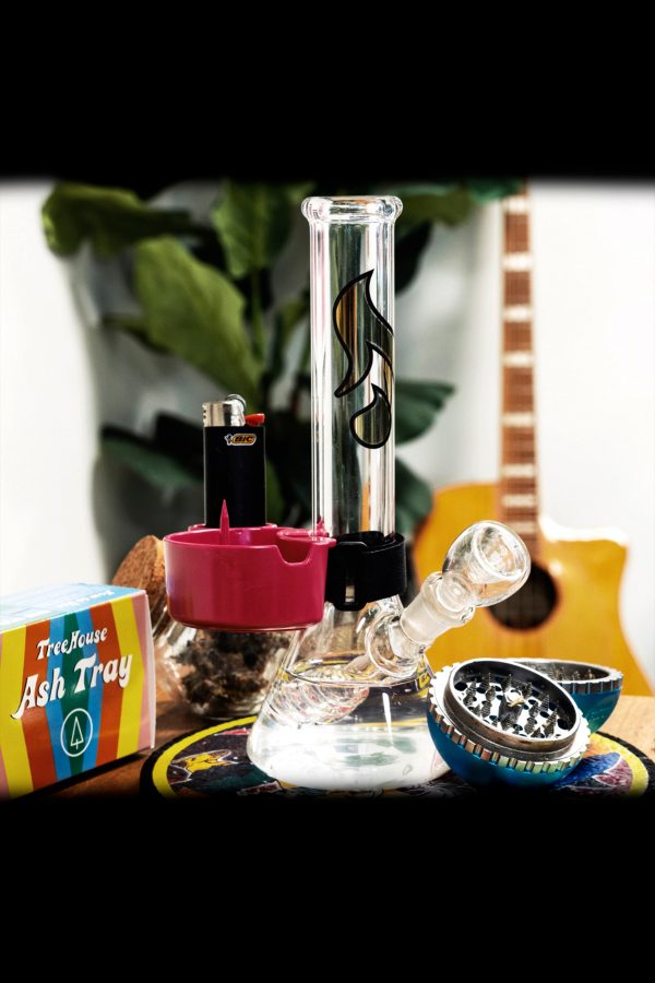 A glass bong with a pink accent piece, lighter, grinder, and ashtray on a patterned surface, with a blurred plant and guitar in the background.