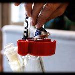 A hand holding a colorful glass cannabis oil rig attachment above a water pipe with a collection tray below.