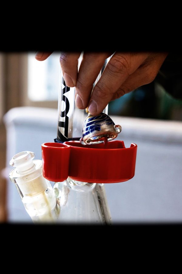 A hand holding a colorful glass cannabis oil rig attachment above a water pipe with a collection tray below.