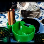 Cannabis accessories including a green grinder, small containers of bud, a lighter, and water bottle arranged on a patterned surface with dramatic lighting.