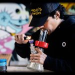 A person using a glass water pipe, illuminated by a lighter flame, while wearing a "Mango Flowers" branded hat.